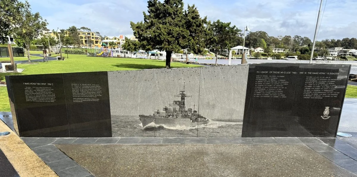 Memorial wall with a central grayscale ship engraving and side text panels in a grassy waterfront park setting.