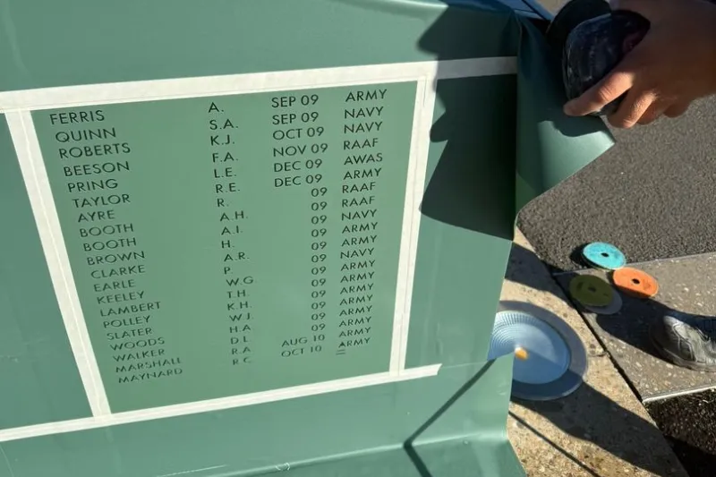 Green memorial panel listing names, initials, dates, and service branches (Army, Navy, RAF); a hand rests at the top edge.