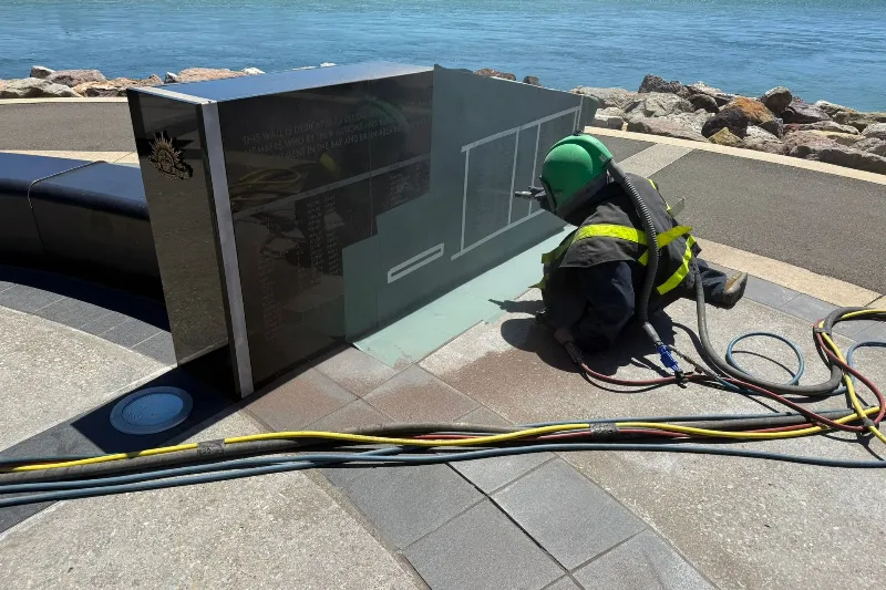 Worker in a green helmet and safety suit kneels beside a glass panel sculpture near the rocky coastline, with cables and hoses on the ground.