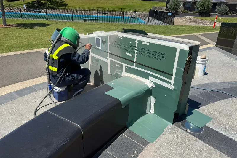 Worker in a safety vest and green helmet power-washes or paints a large green outdoor sculpture with a hose and spray tool.