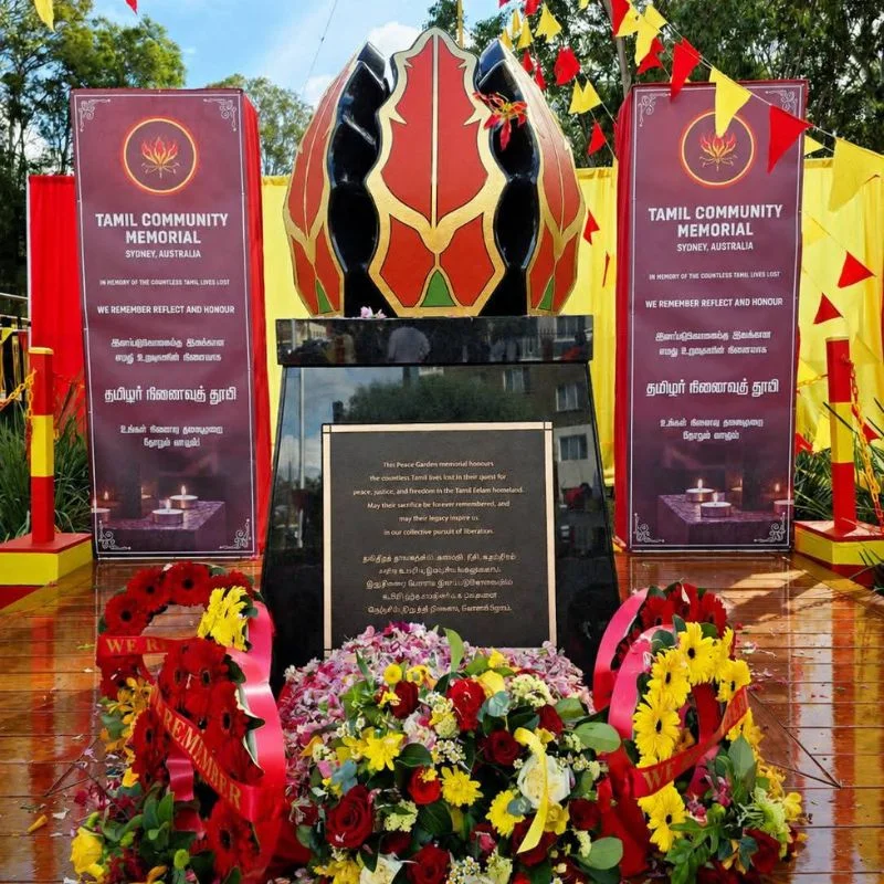 The completed Tamil Community Memorial beautifully decorated for an event. The monument features the painted red and gold flower sculpture atop a polished black pedestal, surrounded by large wreaths of vibrant red and yellow flowers.