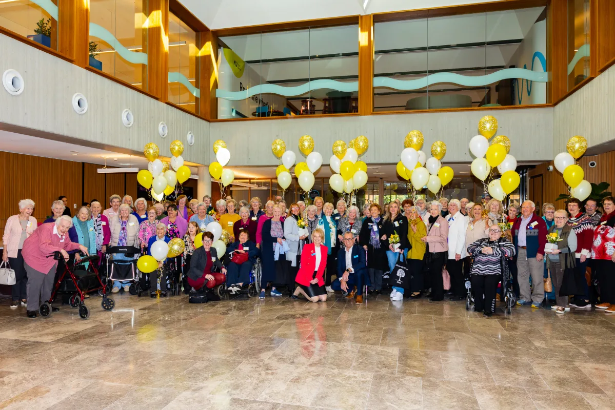 arge group gathered indoors with yellow and white balloons for a community celebration.