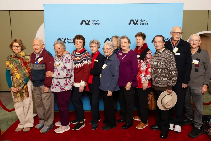 Group of Lantern Club members gathered in front of a NextSense backdrop at a community event.